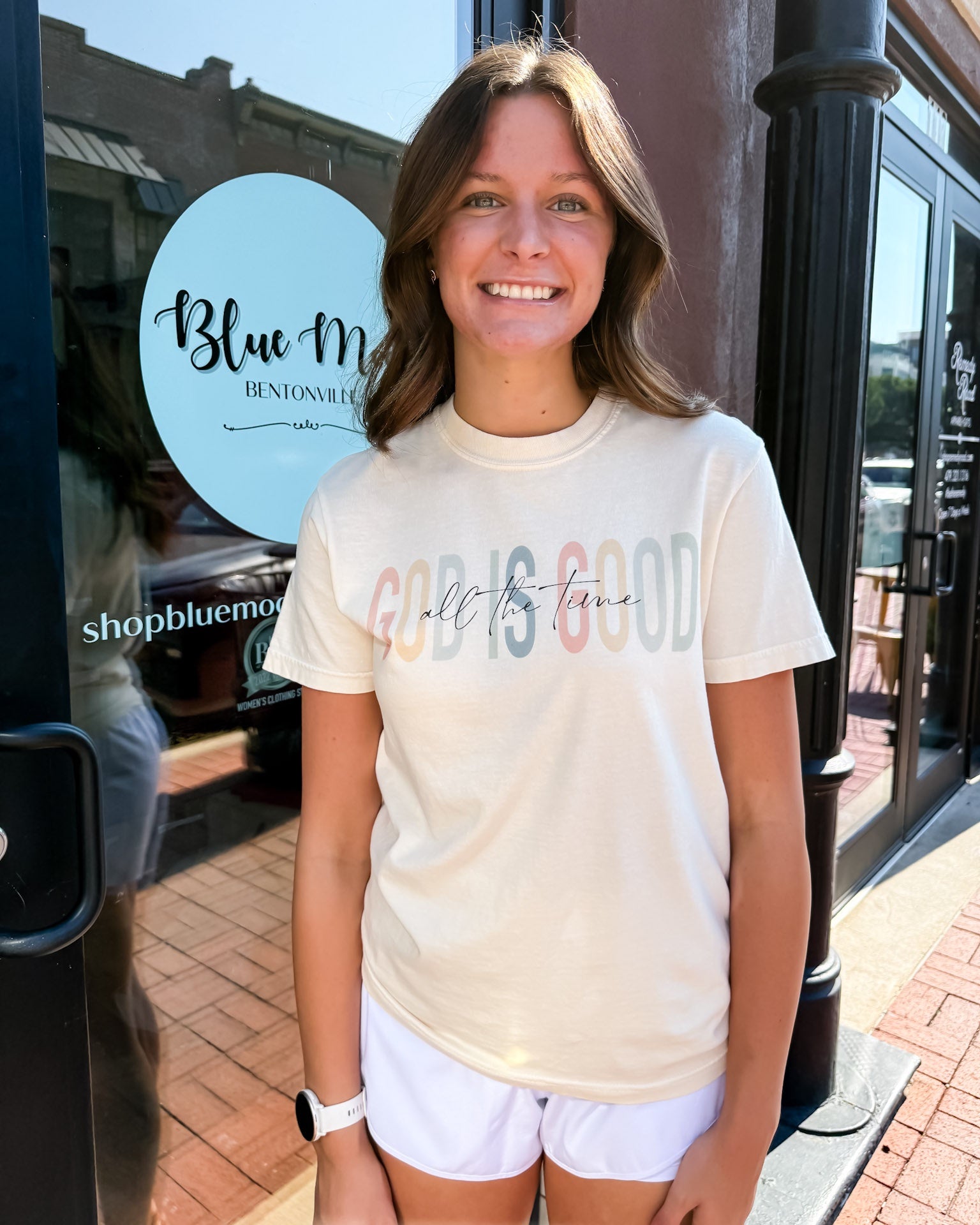 Woman wearing a 'Good is Good' t-shirt in front of a store with a Blue Moon Bendon Hills sign.