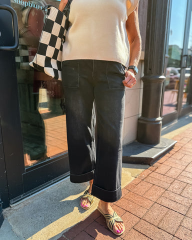 Person wearing a beige top and black pants standing on a sidewalk with a store in the background.