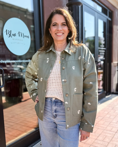 Woman wearing a green jacket with floral patterns in front of a store named 'Blue Moon'.