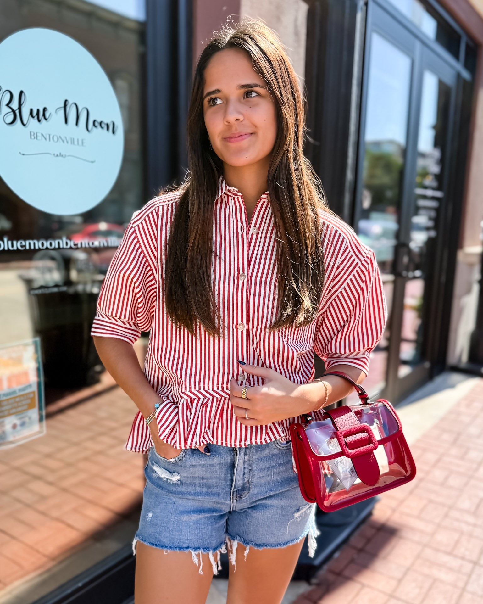 Woman in a red and white striped shirt and denim shorts holding a red handbag in front of a store.