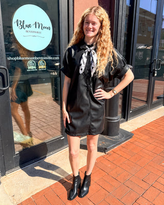 Model wearing a black faux leather shirt dress with cuffed short sleeves and a button-front collar, styled with a checked scarf and black ankle boots