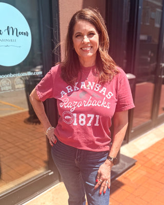 Model wearing a vintage cardinal Arkansas Razorbacks 1871 graphic t-shirt with distressed white lettering, football graphics, a crew neckline, short sleeves, and dark wash denim jeans.