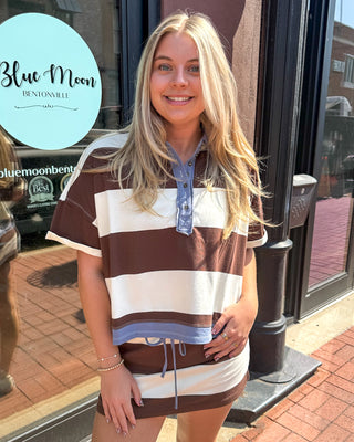 Woman wearing cocoa brown and cream striped polo top with chambray collar, standing outside Blue Moon Bentonville storefront