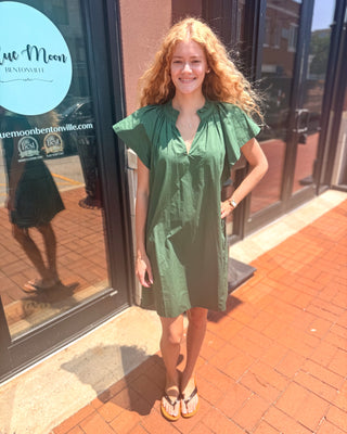 Model wearing a hunter green cotton mini dress with flutter sleeves, a split ruffle neckline, relaxed fit, side pockets, and mini-length hem.