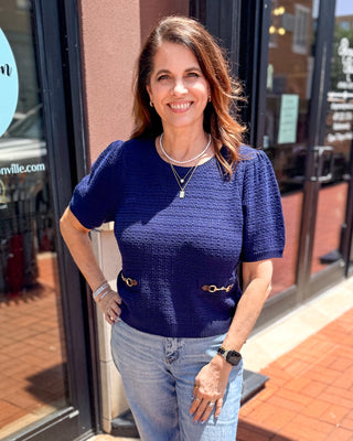 Model wearing a navy textured crewneck sweater with short puff sleeves and decorative gold horsebit waist details paired with light wash denim.
