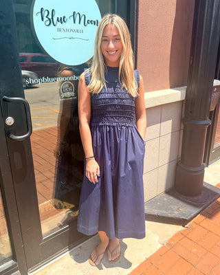 Model wearing a navy sleeveless cotton midi dress with a smocked bodice, ruffle neckline, side pockets, and a flowy skirt paired with flat sandals.