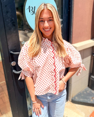Model wearing a red polka dot blouse with scalloped front trim, puff sleeves, tie cuffs, and a stand collar paired with light wash denim.