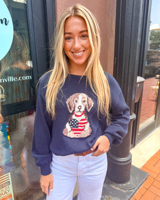 Model wearing a navy knit sweater featuring a puppy graphic holding an American flag heart with long sleeves and a relaxed fit.