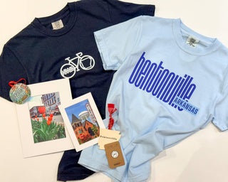 Two t-shirts, one black with a bicycle graphic and one light blue with 'Bentonville Arkansas' text, alongside postcards and a small bag.