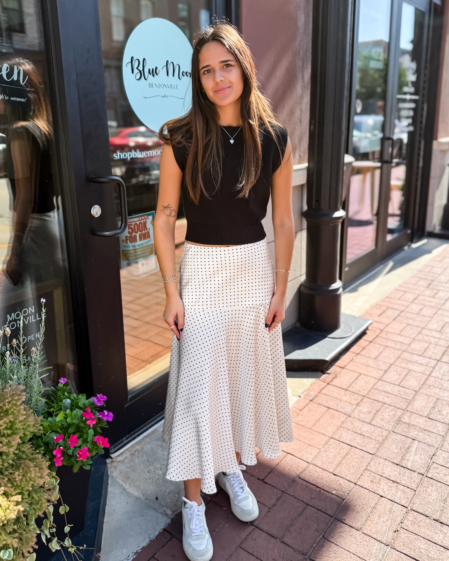Woman in a black top and white patterned skirt standing on a sidewalk with storefronts in the background.