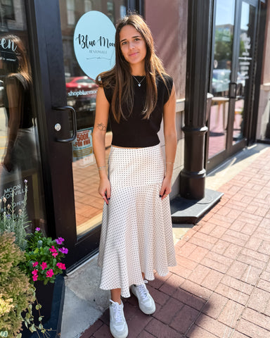 Woman in a black top and white patterned skirt standing on a sidewalk with storefronts in the background.