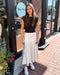Woman in a black top and white patterned skirt standing on a sidewalk with storefronts in the background.