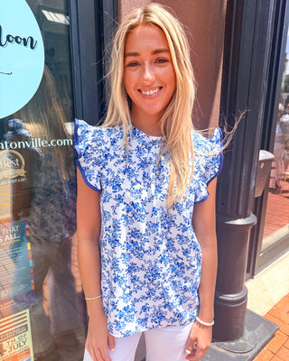 Model wearing a blue and white floral blouse with ruffle cap sleeves and gathered neckline paired with white pants.