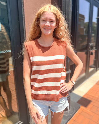 Model wearing a caramel and ivory striped sleeveless sweater vest with a crew neckline, decorative gold side buttons, ribbed hem, and relaxed fit paired with light wash denim shorts.