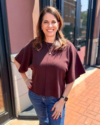 Model wearing a chocolate brown lightweight knit sweater with a crew neckline, elbow-length wide sleeves, relaxed fit, and center front seam paired with blue jeans.