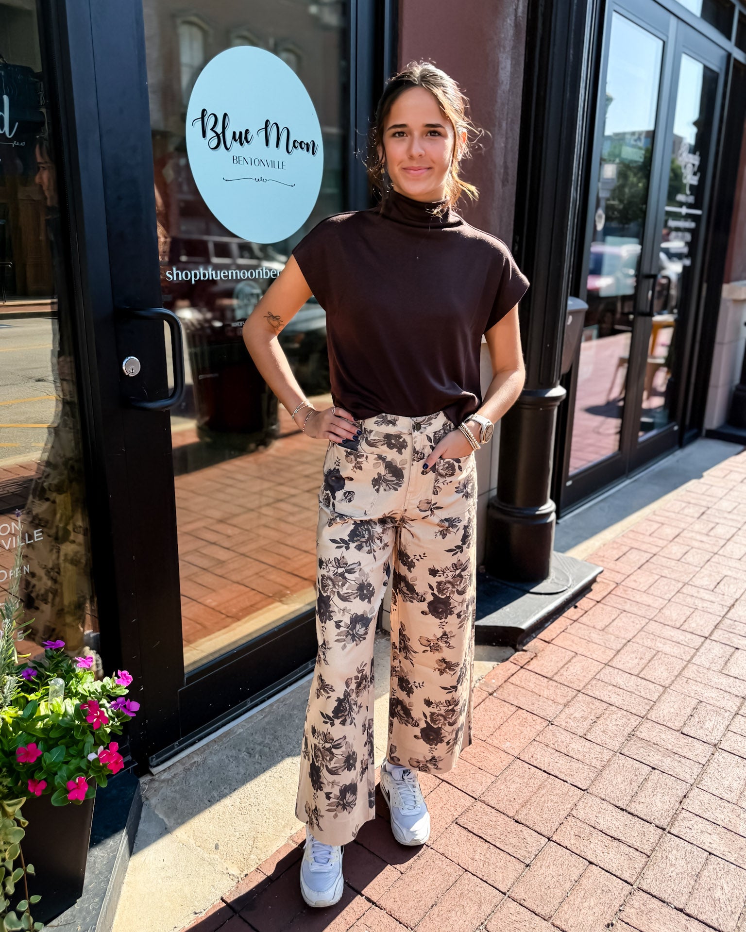 Woman standing outside a store named 'Blue Moon' wearing a brown top and floral pants.