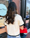 Woman with long hair wearing a white top with black trim and blue jeans, holding a red handbag, standing outside a store.