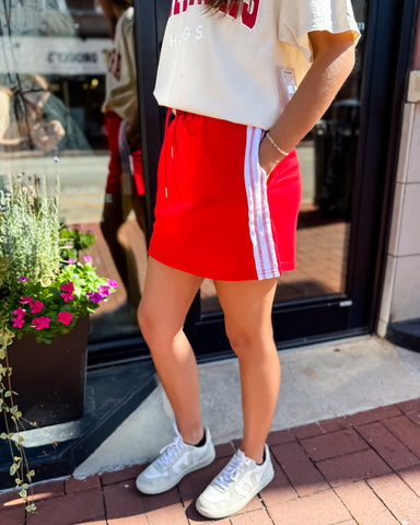 Person wearing a red skirt with white stripes and a white top, standing on a sidewalk.