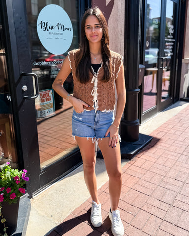 Woman standing outside a building with 'Blue Moon' branding, wearing a brown crochet top and denim shorts.