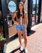 Woman standing outside a building with 'Blue Moon' branding, wearing a brown crochet top and denim shorts.