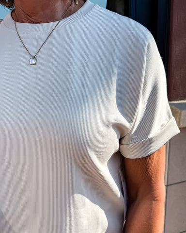 Person wearing a light-colored ribbed shirt with a necklace, standing outdoors.