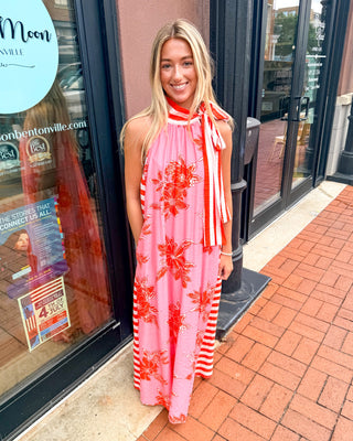Model wearing a pink midi dress with orange botanical floral print, striped scarf-tie halter neckline, and flowy summer silhouette.