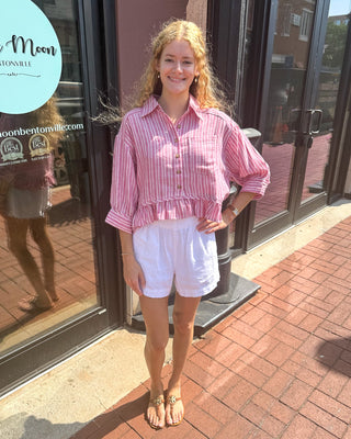 Woman wearing raspberry and white striped button-down ruffled hem top with white shorts, standing outside Blue Moon Bentonville storefront