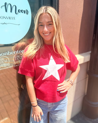 Model wearing a red lightweight mock neck sweater with an oversized white star graphic, short sleeves, ribbed trim, and a relaxed fit paired with light wash denim.