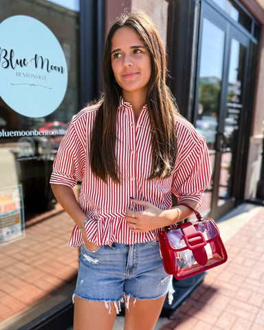 Woman in a red and white striped shirt and denim shorts holding a red handbag in front of a store.