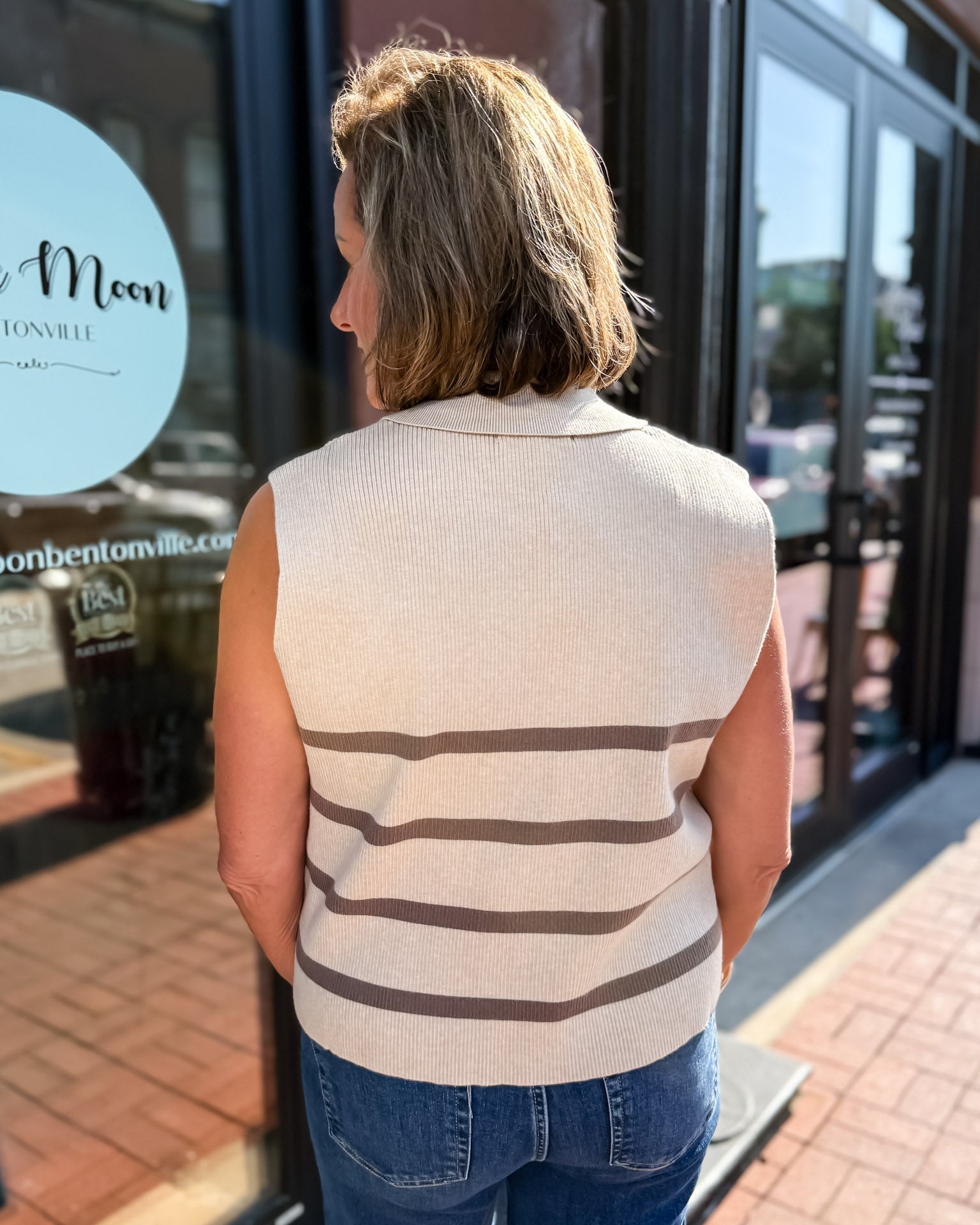 Woman wearing a striped vest and jeans standing in front of a store entrance.