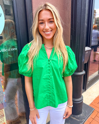 Model wearing a summer green cotton blouse with a ruffled collar, covered button front, and puff sleeves paired with white pants.