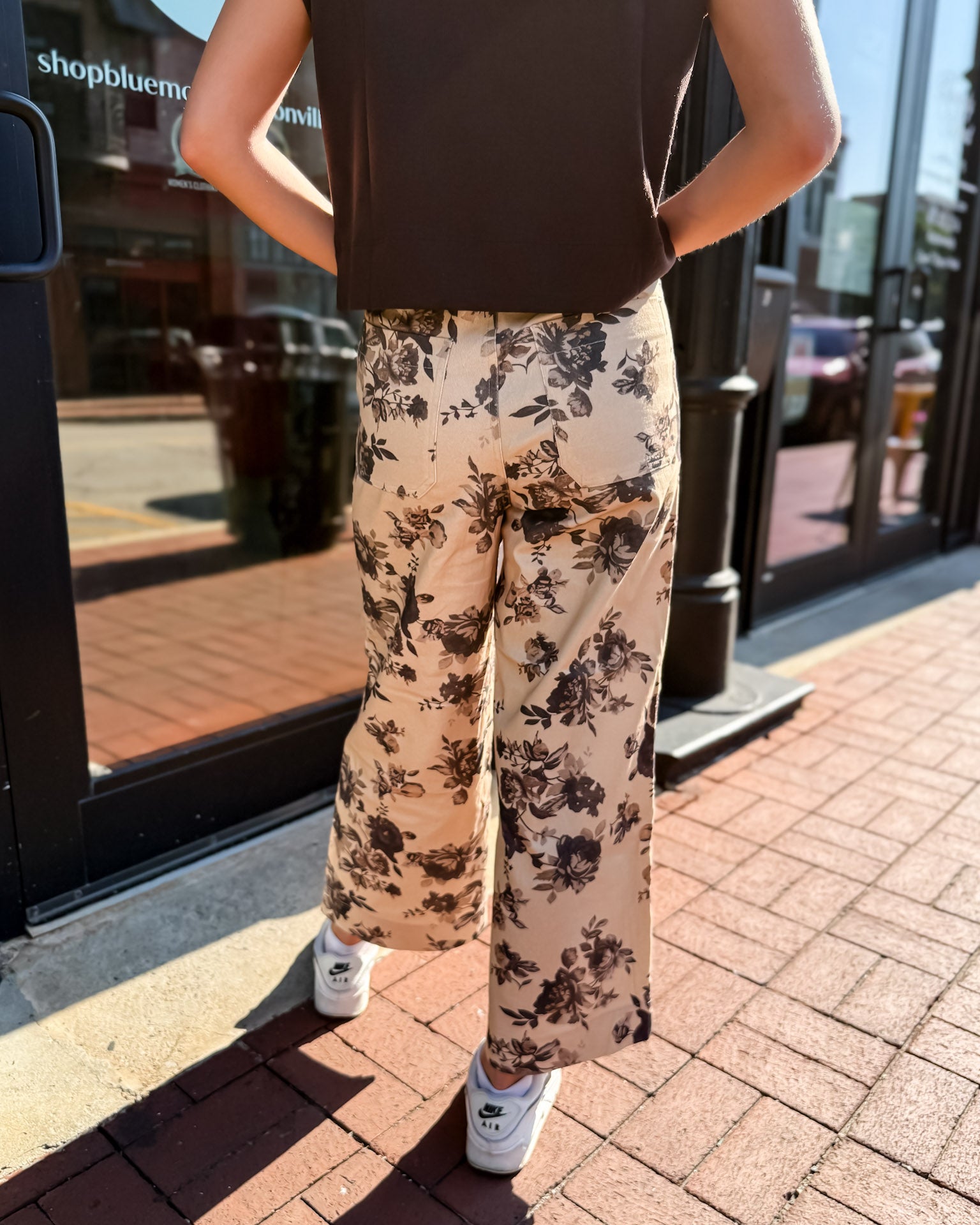 Person wearing floral pants and a dark top standing on a sidewalk.