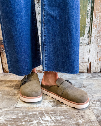 Person wearing olive green suede shoes with white soles on a wooden floor.