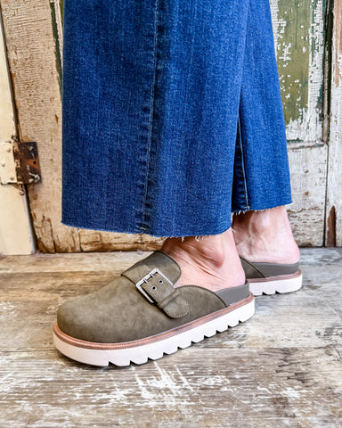 Person wearing olive green suede shoes with white soles on a wooden floor.