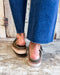 Olive green sandals worn with blue jeans on a wooden floor.