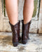 Brown cowboy boots worn by a person on a wooden floor with a rustic background